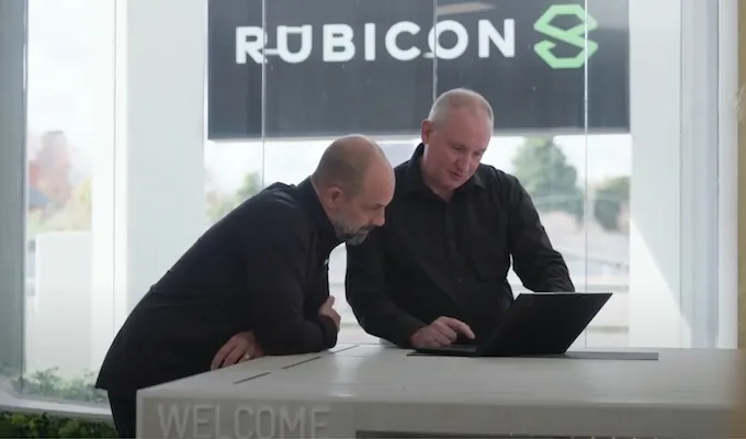 Two men at a counter, one using a laptop, with a 'Rubicon 8' logo in the background