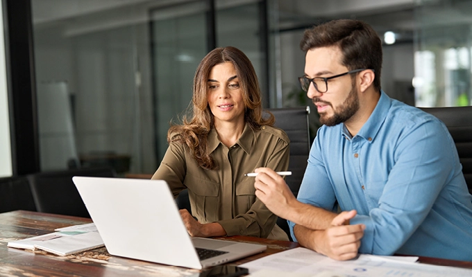 Two busy colleagues working together talking using laptop in office